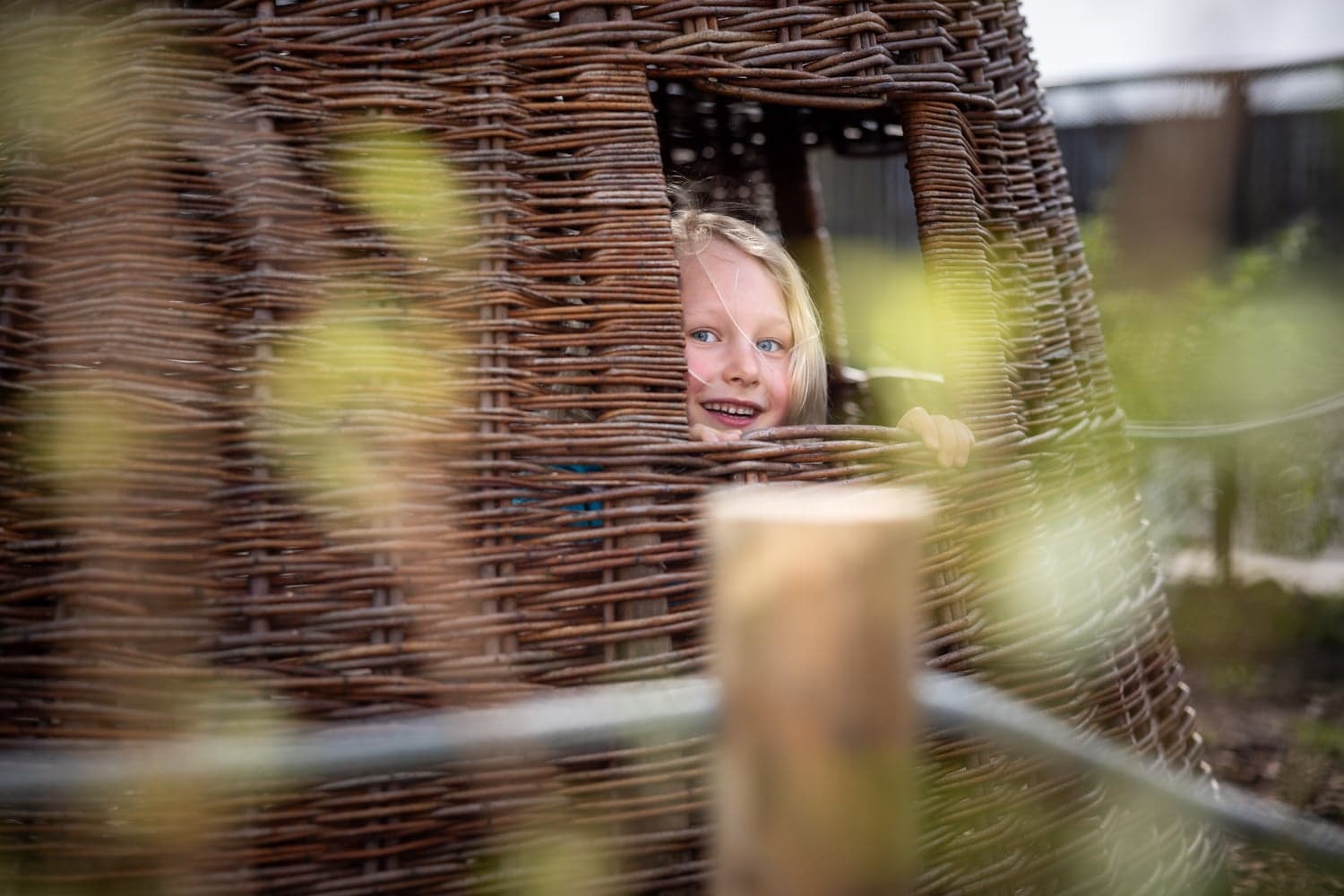 Blij kind kijkt in tuin door rieten huisje bij kinderopvang Kind&co ludens