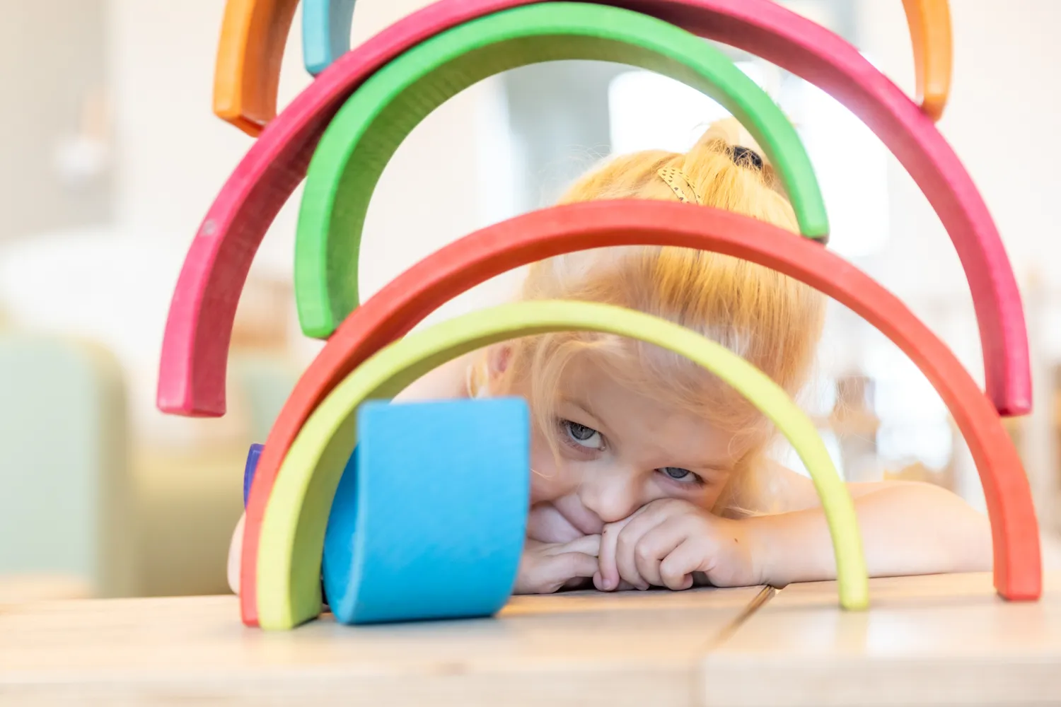 Kind kijkt door gestapelde houten regenboog bij kinderopvang Kind&co ludens
