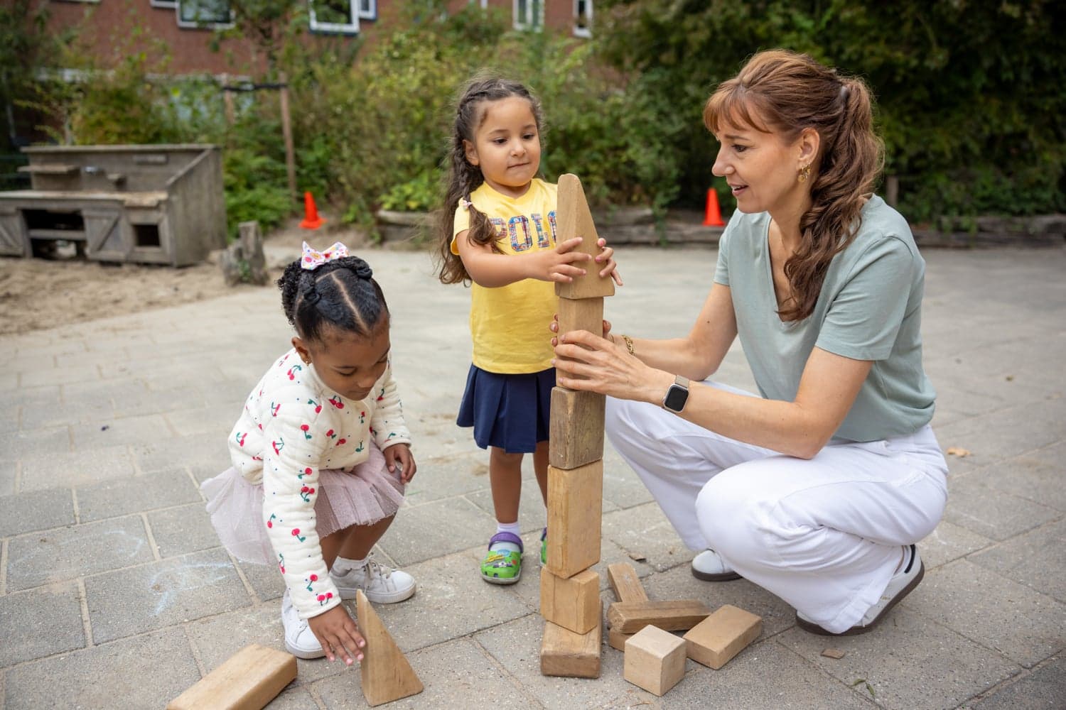Pedagogisch medewerker bouwt buiten een houten blokkentoren met twee kinderen bij kinderopvang Kind&co ludens