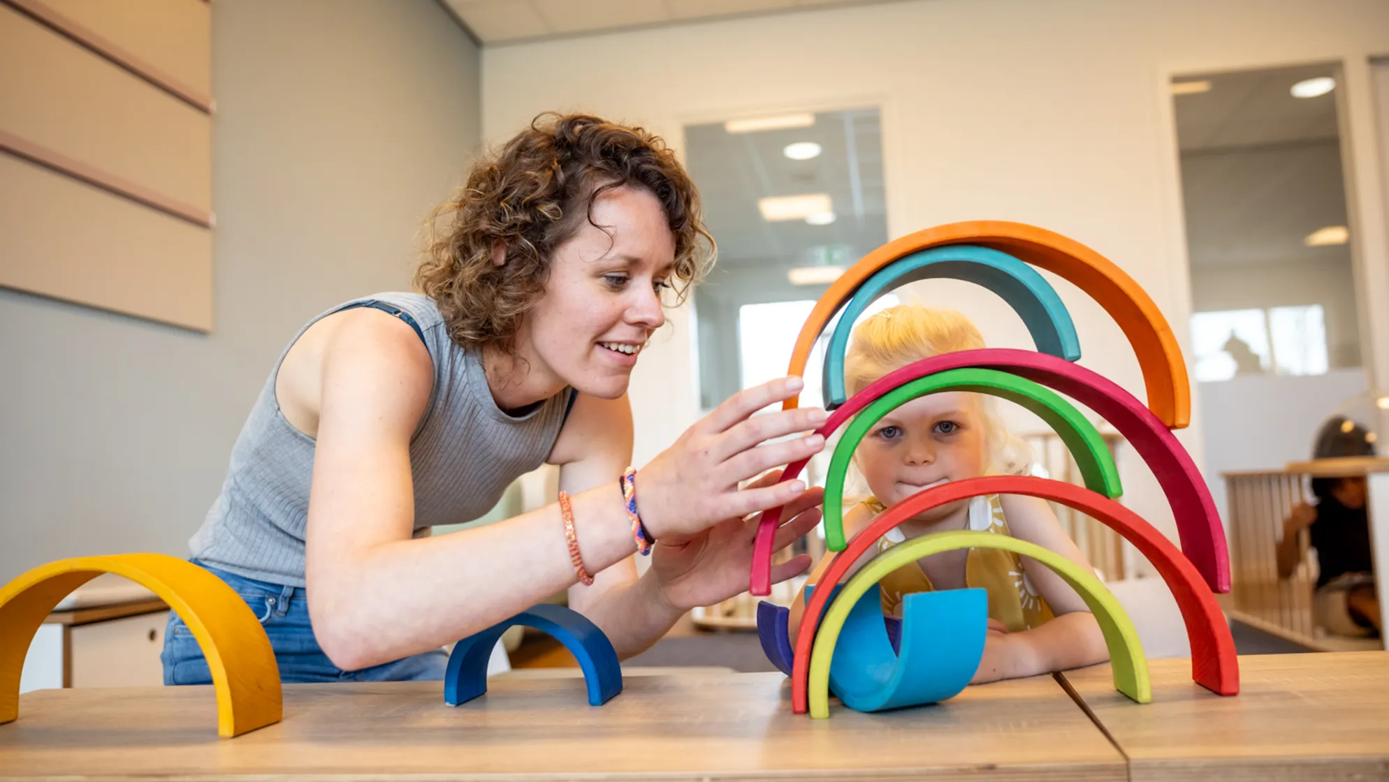 Kind stapelt houten regenboog samen met pedagogisch medewerker bij kinderopvang Kind&co ludens