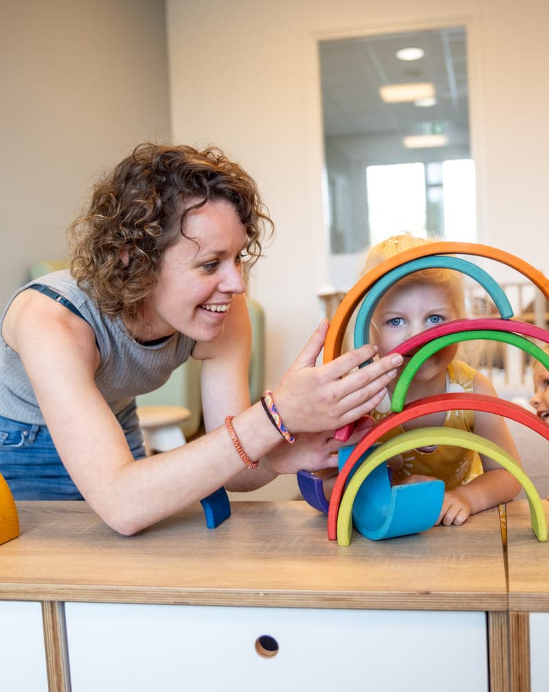 Kind stapelt houten regenboog samen met pedagogisch medewerker bij kinderopvang Kind&co ludens