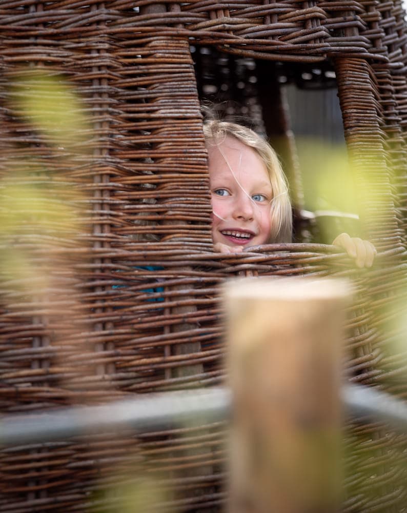 Blij kind kijkt in tuin door rieten huisje bij kinderopvang Kind&co ludens