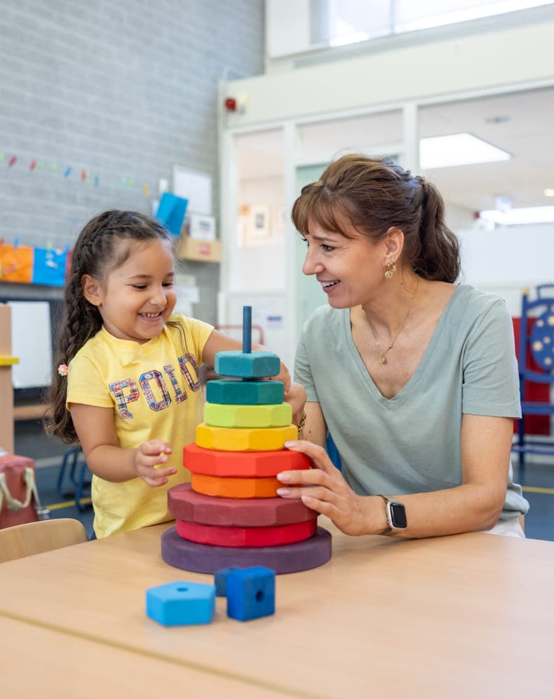 Kind stapelt houten ringen samen met pedagogisch medewerker bij kinderopvang Kind&co ludens