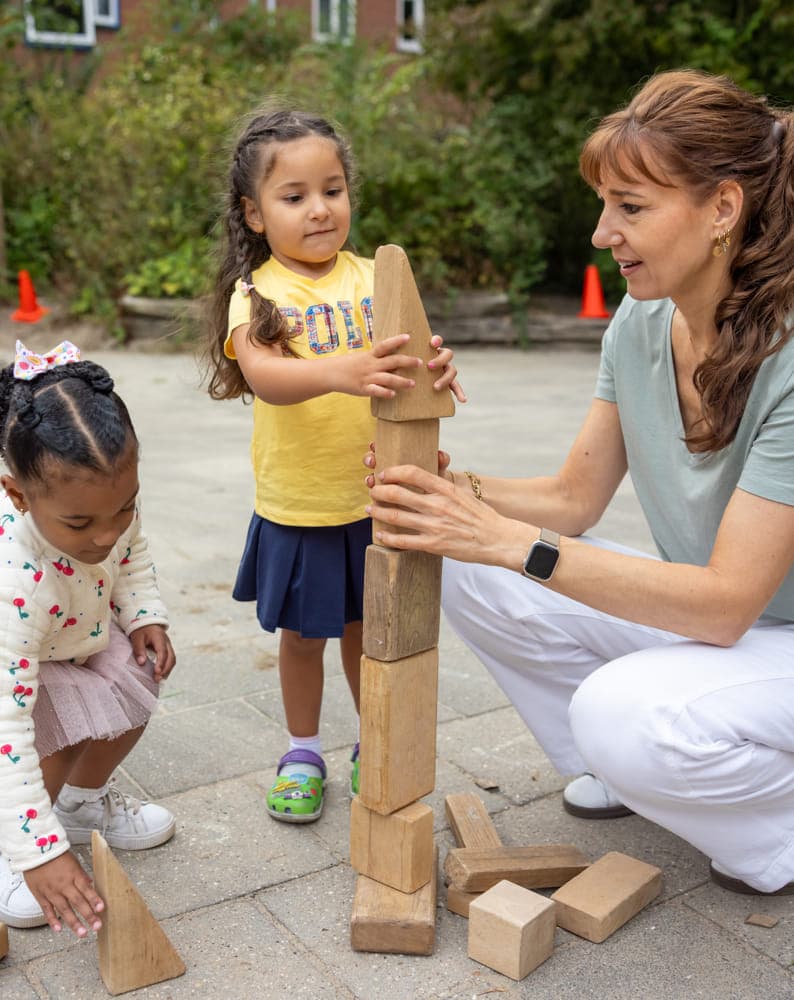 Pedagogisch medewerker bouwt buiten een houten blokkentoren met twee kinderen bij kinderopvang Kind&co ludens