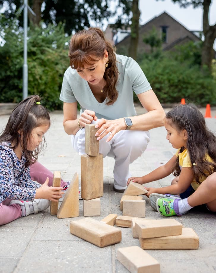 Pedagogisch medewerker bouwt buiten een houten blokkentoren met twee kinderen bij kinderopvang Kind&co ludens