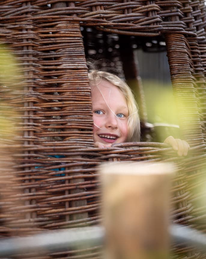 Blij kind kijkt in tuin door rieten huisje bij kinderopvang Kind&co ludens