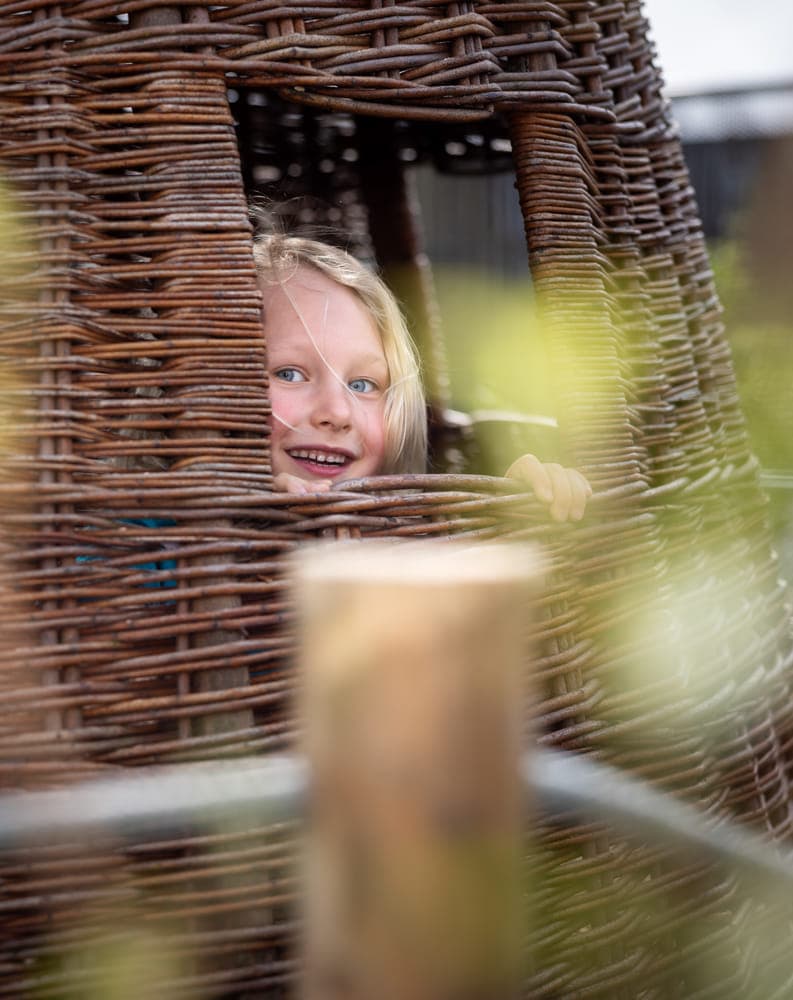 Blij kind kijkt in tuin door rieten huisje bij kinderopvang Kind&co ludens