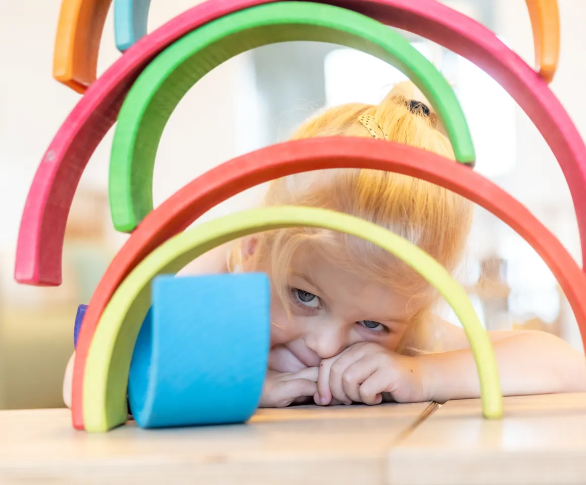 Kind kijkt door gestapelde houten regenboog bij kinderopvang Kind&co ludens