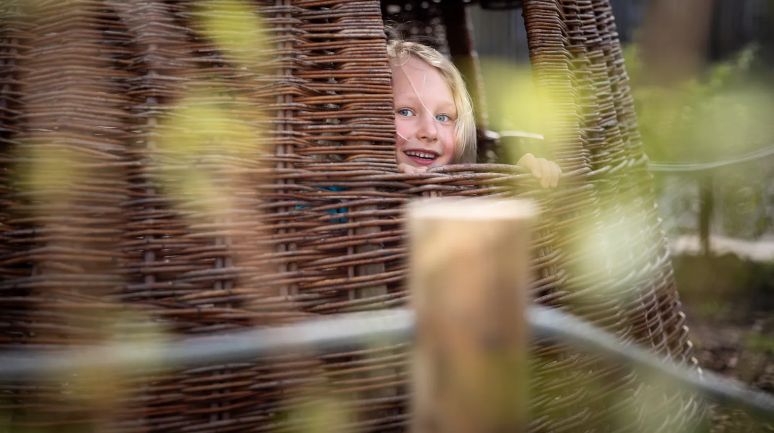 Blij kind kijkt in tuin door rieten huisje bij kinderopvang Kind&co ludens
