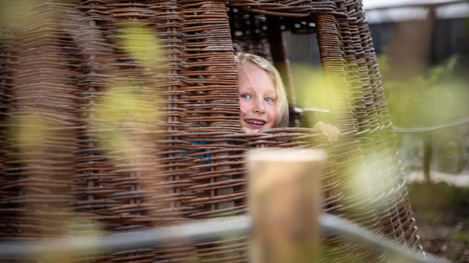 Blij kind kijkt in tuin door rieten huisje bij kinderopvang Kind&co ludens